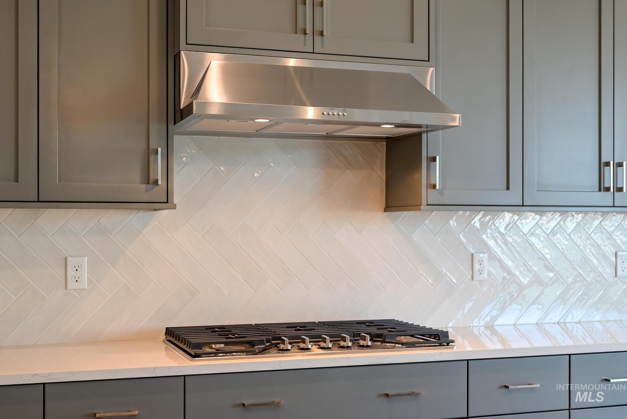 Kitchen featuring gray cabinetry, under cabinet range hood, backsplash, light stone counters, and stainless steel gas cooktop