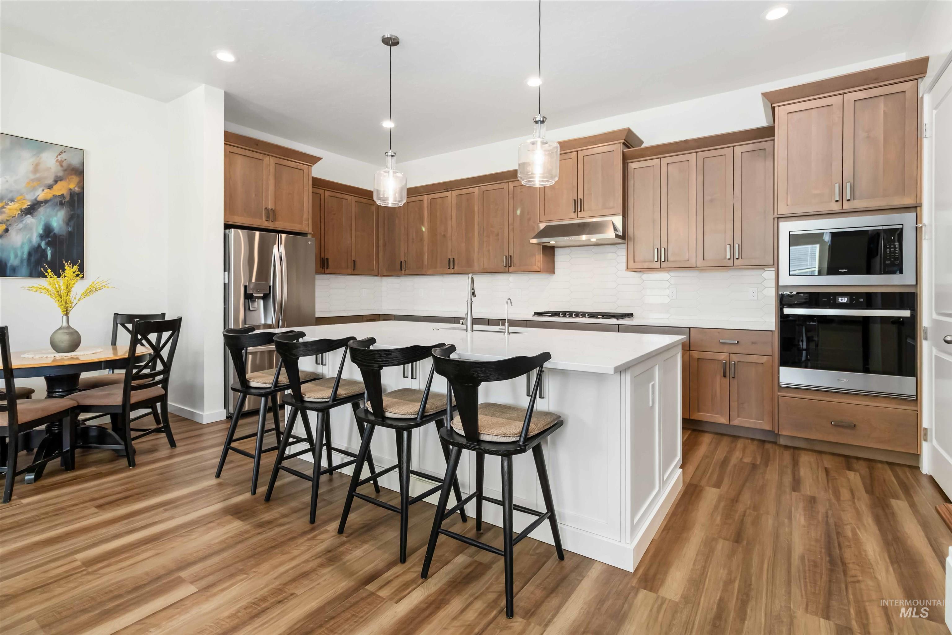 Kitchen featuring large oversized island with sink, quartz countertops neutral backsplash, stainless steel appliances, hanging modern farm style light fixtures.
