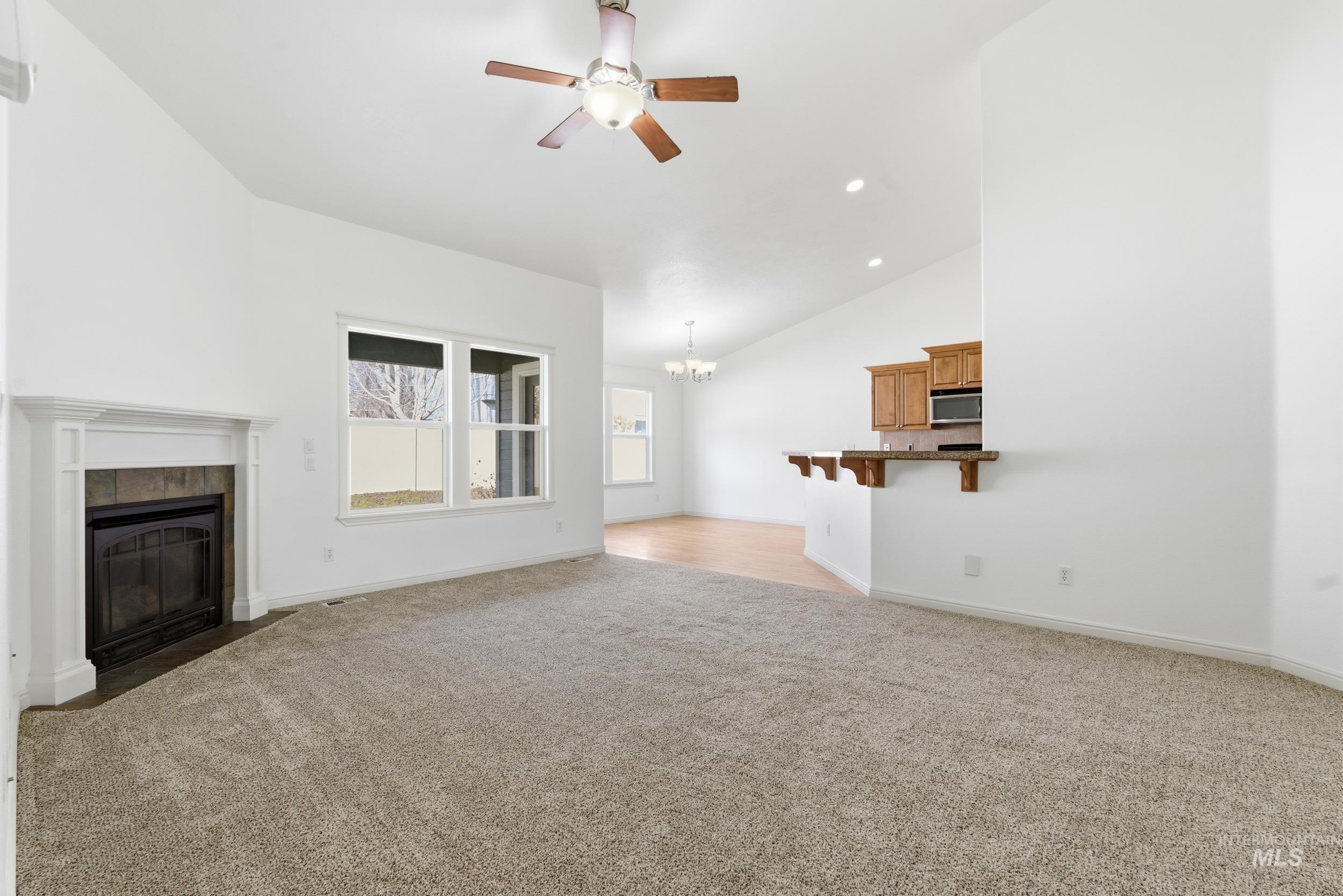 Unfurnished living room featuring a tiled fireplace, light carpet, a ceiling fan, lofted ceiling, and a chandelier