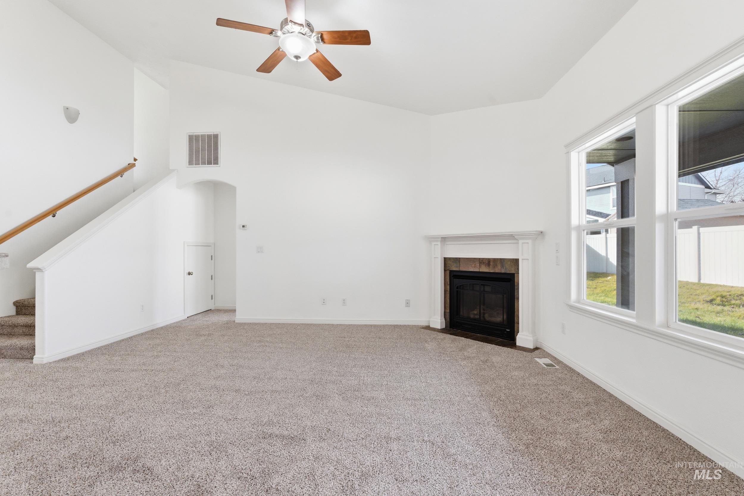 Unfurnished living room featuring stairs, light colored carpet, high vaulted ceiling, a ceiling fan, and a tile fireplace