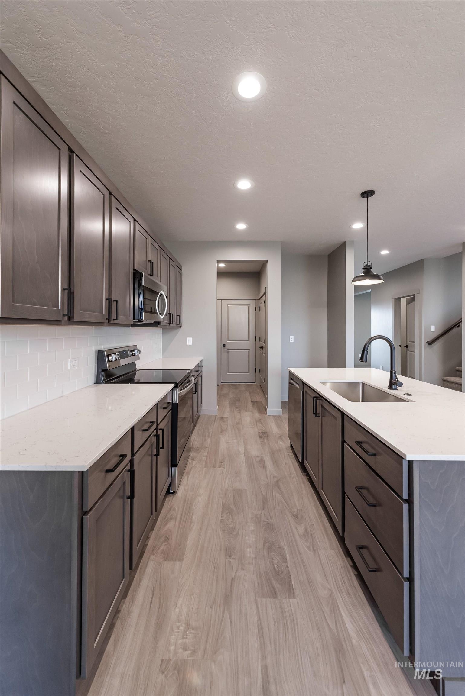 Kitchen with appliances with stainless steel finishes, pendant lighting, light stone counters, light wood-style flooring, and a kitchen island with sink
